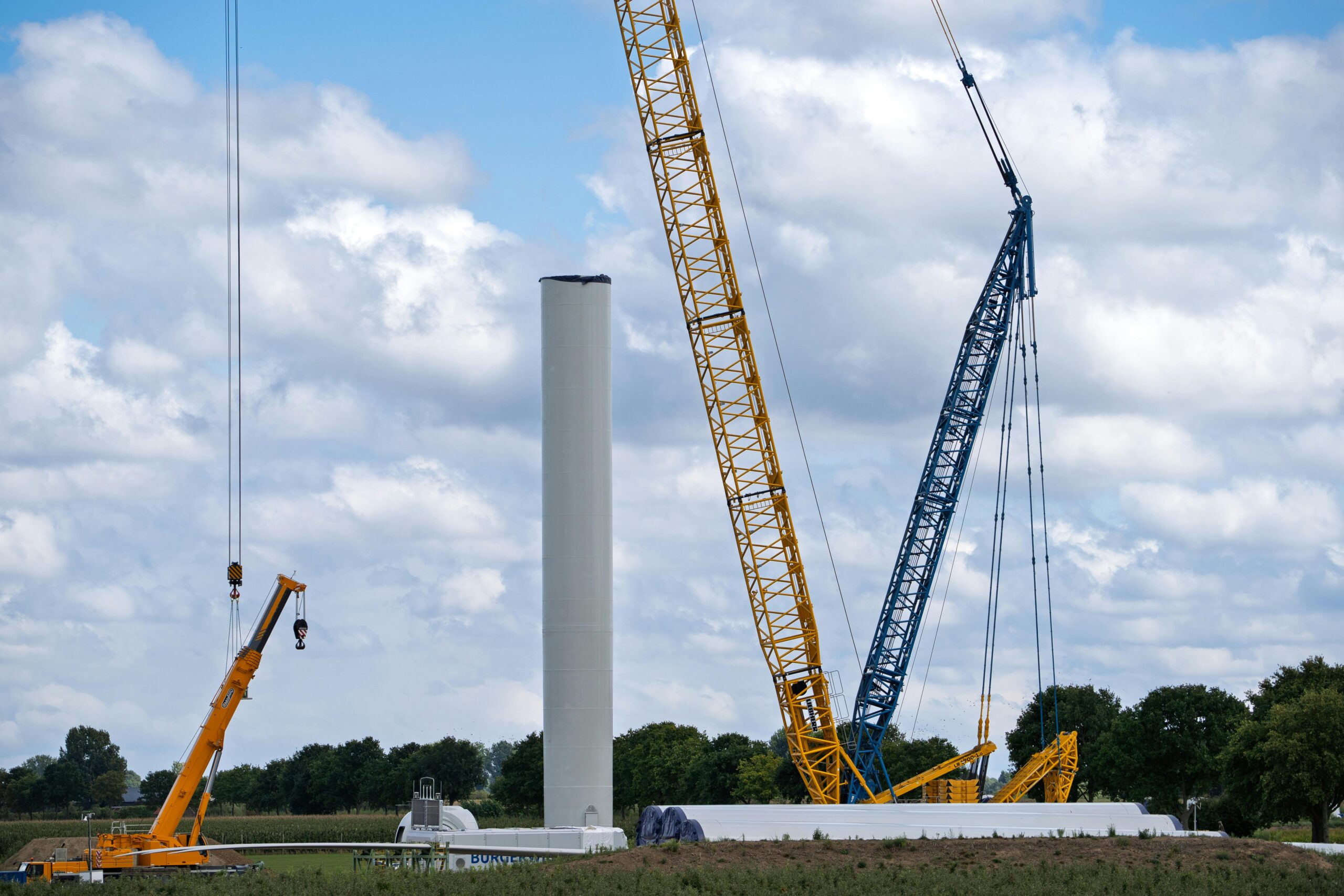 Cranes assembling a wind turbine tower in an open field under a cloudy sky.